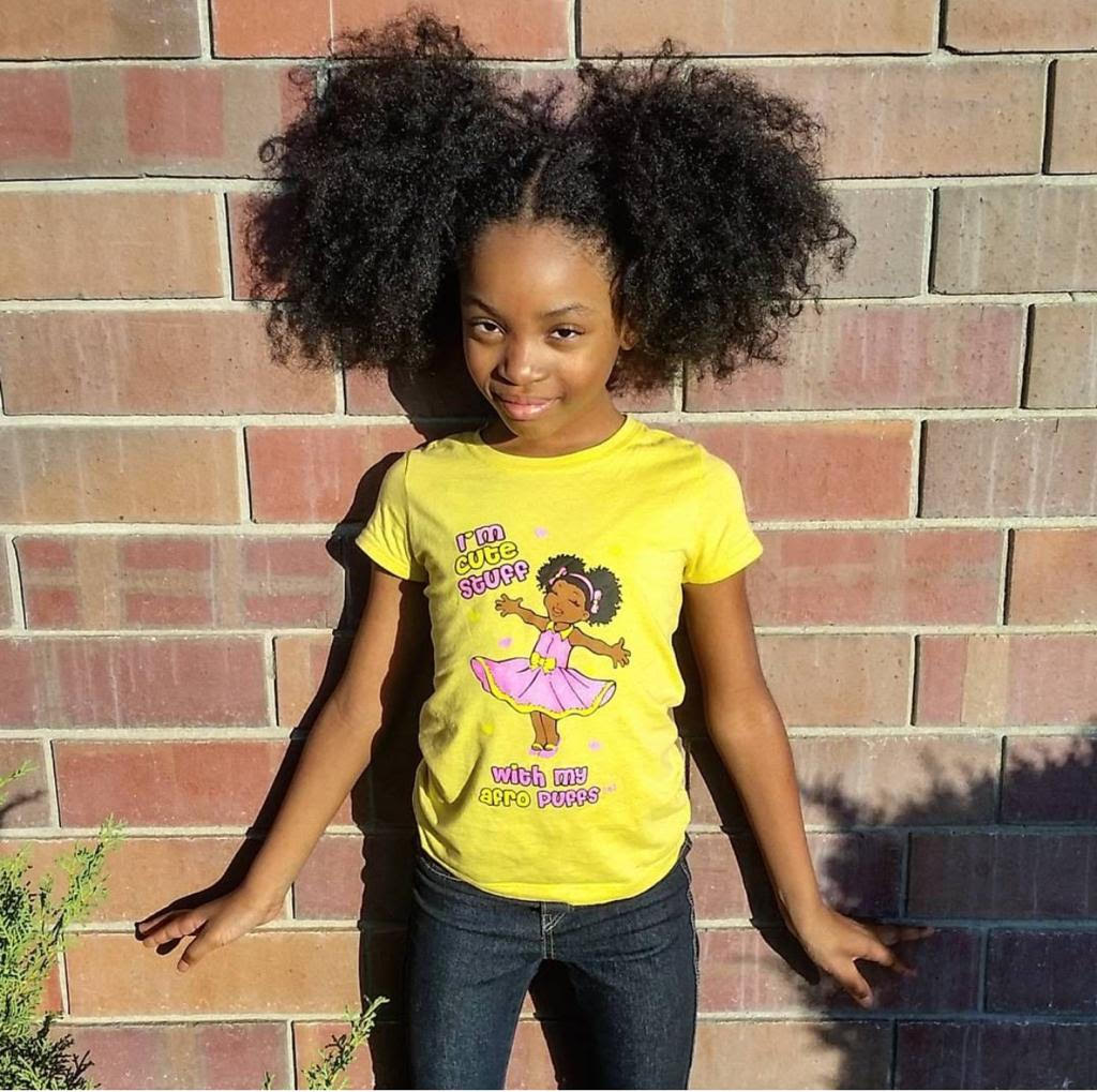 Young girl wearing a yellow t-shirt with a graphic design in front of a brick wall.